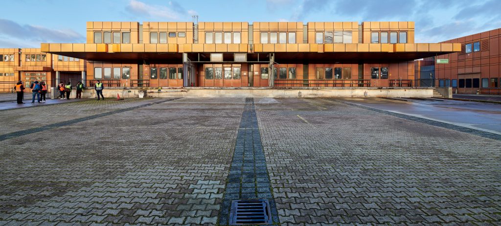 Auf dem Bild ist die Frachtkantine auf dem ehemaligen Gelände des Flughafen Tegel in Berlin-Reinickendorf zu sehen. Die Sonne scheint auf das bronze-farbene Gebäude aus den 1970ern. Davor ist eine große gepflasterte Freifläche mit grauen Steinen.